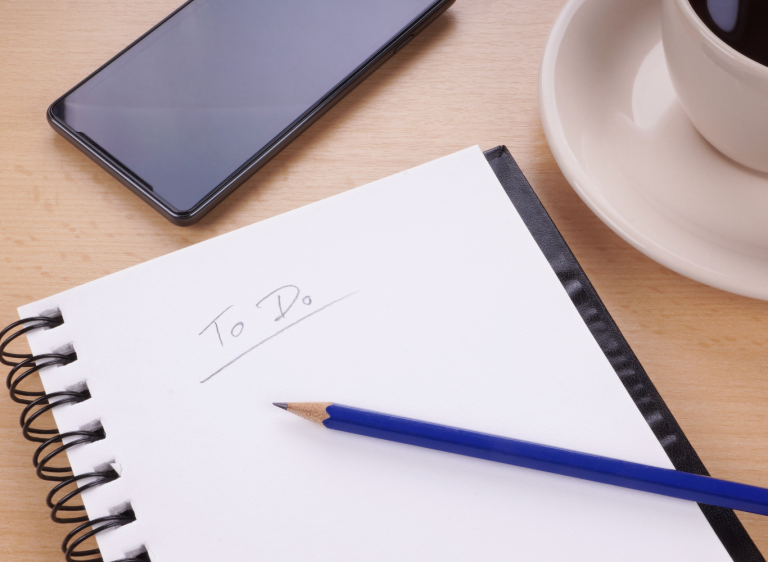 Paper to-do list on desk with pencil, iPhone and coffee.