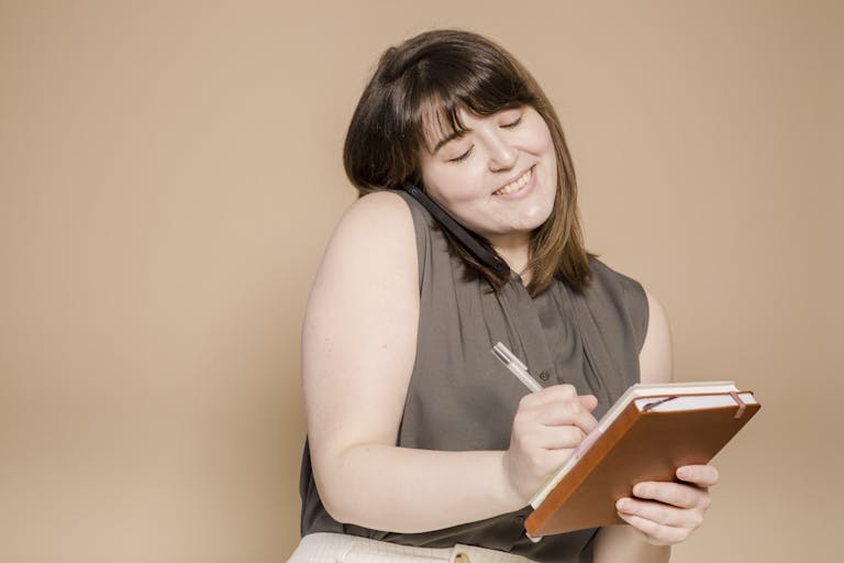Cheerful woman on phone taking notes in a beige studio setting.