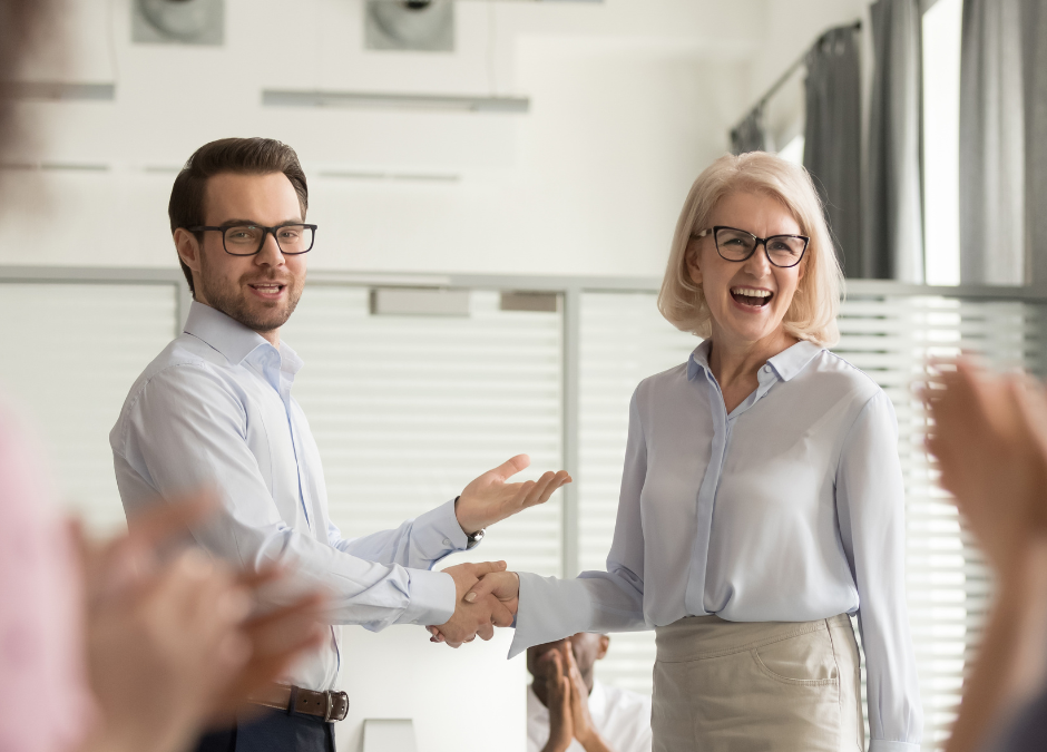 Man shaking hands with smiling woman as coworkers clap and celebrate her on administrative professionals' day.