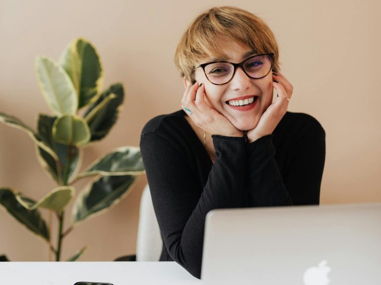 Cheerful woman in eyeglasses and black sweater smiling while working at a desk with a laptop indoors as new executive assistant