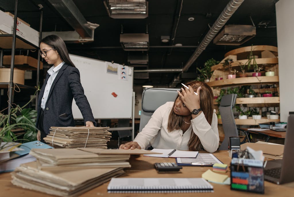 A woman working with an executive assistant on a stack of documents in a modern office setting, one sitting at the desk, the other standing.